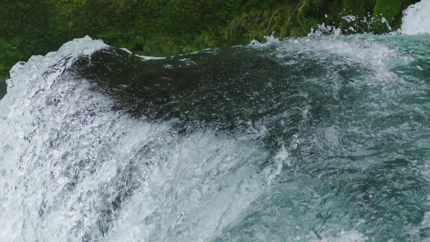The camera pans left at the same speed of the cascading water as it splashes over the lip of a large boulder in the middle of the clean and fresh whitewater river