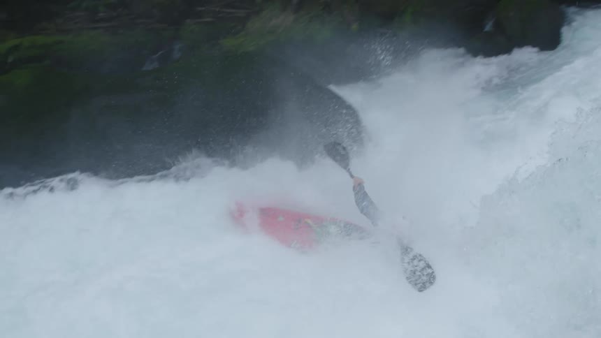 Slow motion reveal of a young man shredding in a whitewater kayak descending a medium size waterfall with green moss and sparkling water