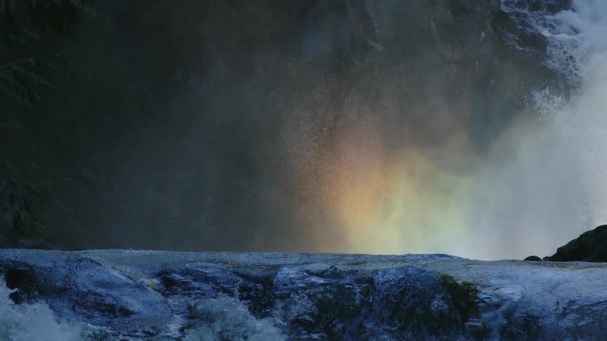 Rainbow forms on the water mist at the base of a waterfall in a whitewater river as the camera slowly tilts down revealing the entire scene