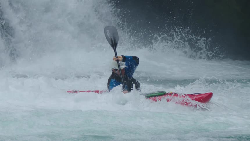 Slow motion shot of a man in a red whitewater kayak emerging from underwater into the air below a big waterfall