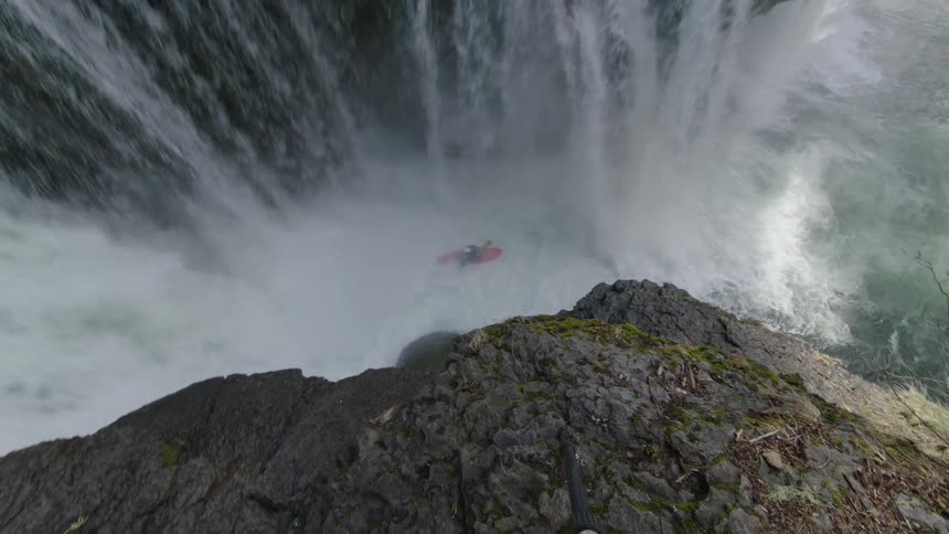 Superwide shot of a man standing at the edge of a cliff with a rope wrapped around him next to a river watches another man in a kayak paddle off of a waterfall and down stream