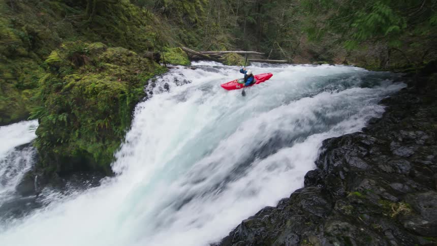 Realtime handheld shot of a man paddling over the lip of a large white waterfall deep in a moss and fern covered gorge in the Pacific Northwest