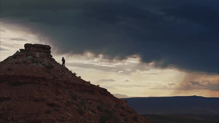 A person climbs to the top of a desert mountain under a stormy sky