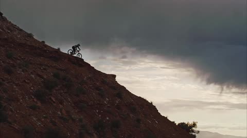 A person on a bike riding down a ridge with the stormy sky and sunset in the background