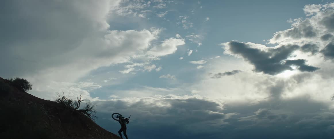 A person is carrying a mountain bike up a hill under a cloudy desert sky