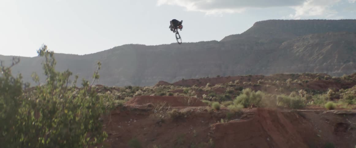 A mountain biker spins a 360 table trick off a desert jump with canyons and mesas