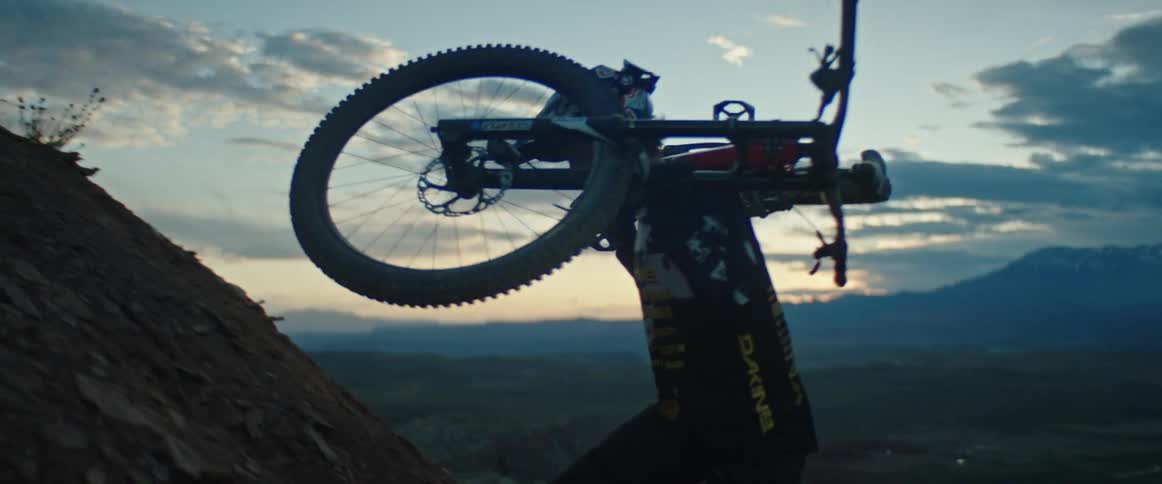 Man carries bicycle up steep slope with desert valley and mountains on horizon at dusk