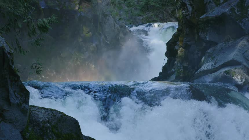 Static shot of two waterfalls pouring over smooth river rock creating large plumes of mist spraying into the sky