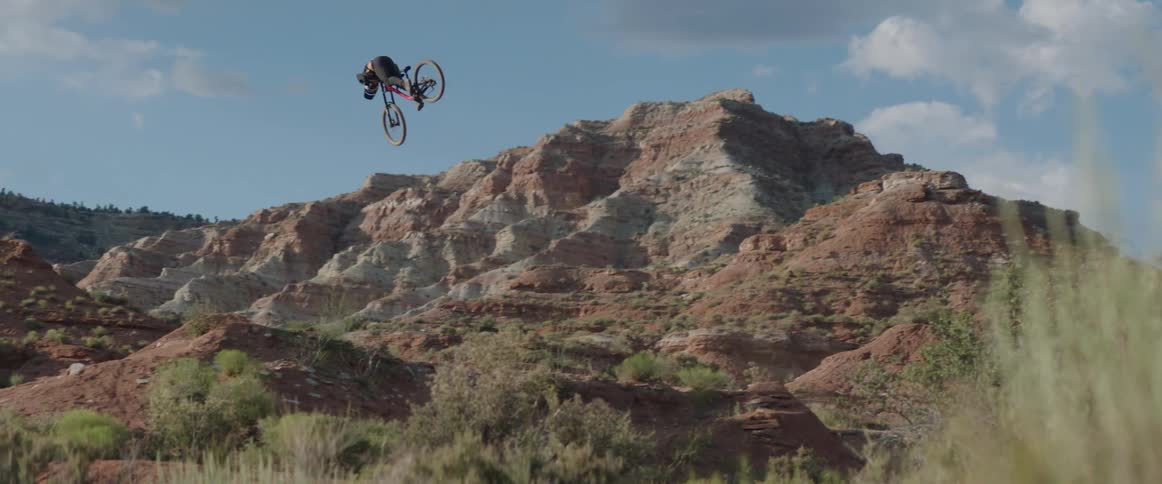 A cyclist on a mountain bike performs a spin trick in the air above a hilly desert in the afternoon