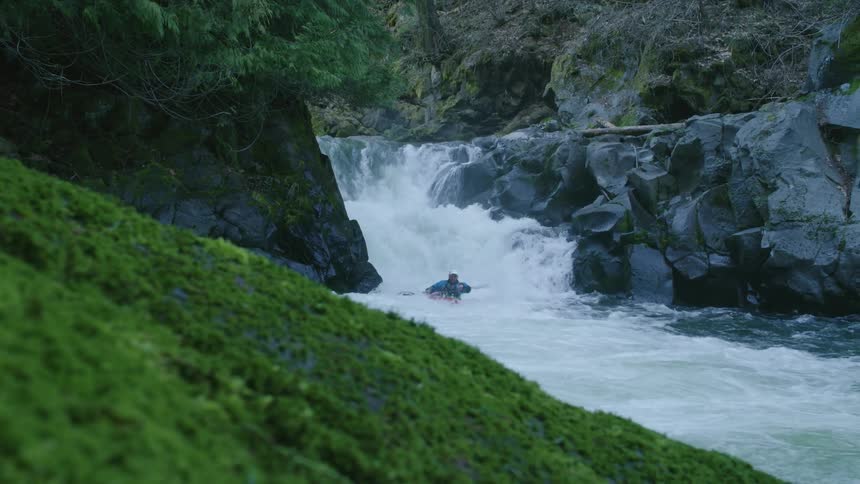 Real time shot with a mossy green foreground of a man in a red whitewater kayak descending a double drop waterfall in this high energy shot