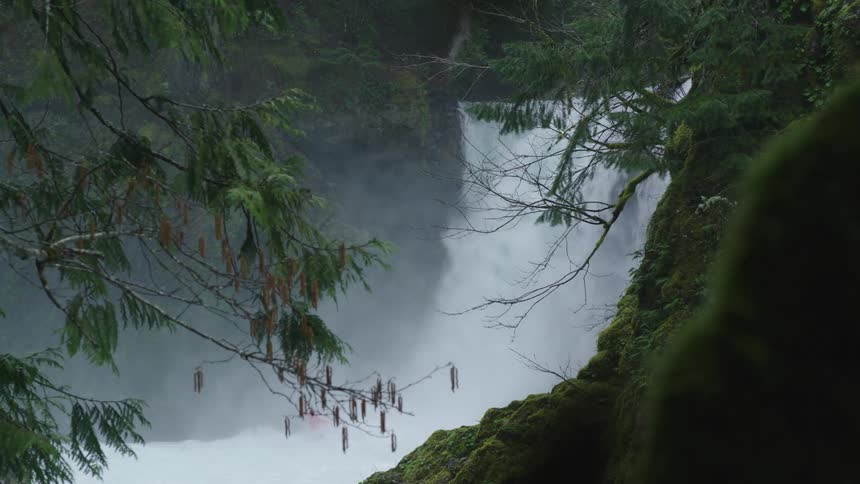 Static shot of a man paddling over a large whitewater waterfall in a moss covered canyon deep in the Pacific Northwest