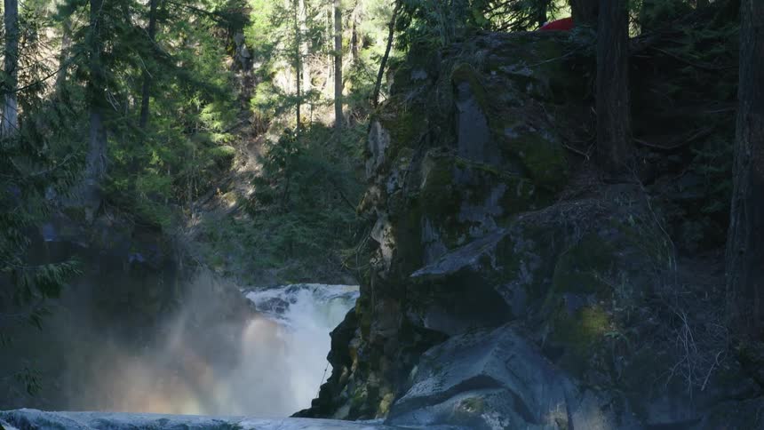 A man portaging his red whitewater kayak next to two beautiful waterfalls with the mist making a stunning rainbow in a deep river gorge