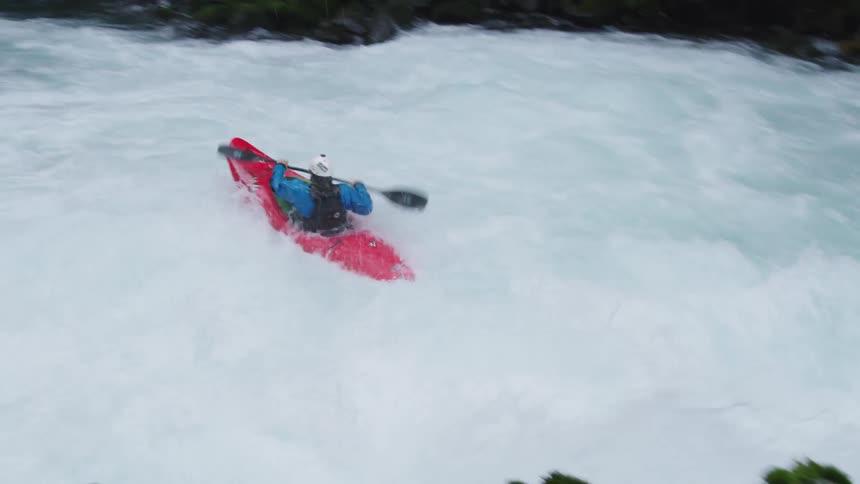 A man in a red whitewater kayak rapidly blasts by the camera as he paddles over a waterfall and sprints downstream