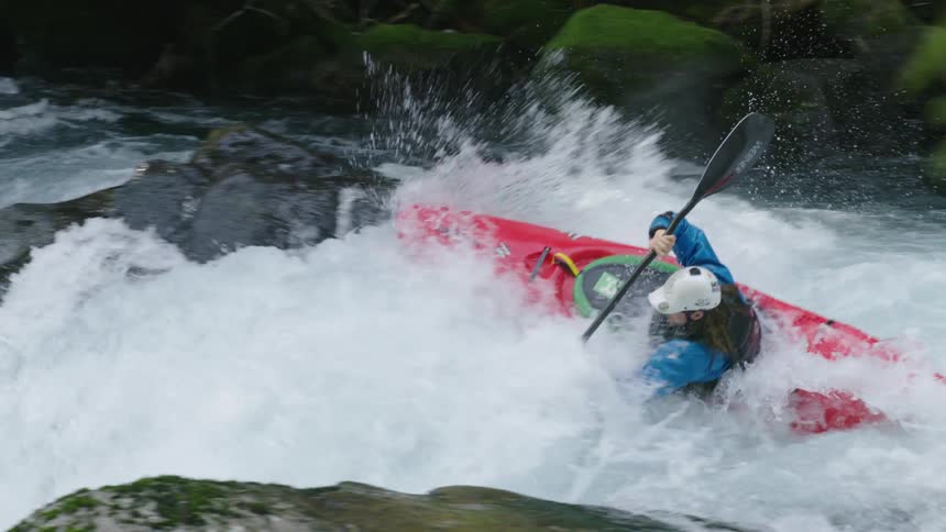 Slow motion shot of a man in a red whitewater kayak launching off rocks and turbulent whitewater before disappearing behind a green moss covered rock