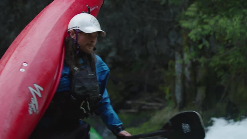 A portaging with a red whitewater kayak ascends a steep cliff with steep raging water behind him