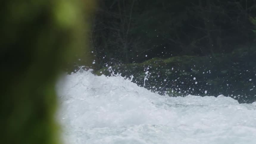 The camera moves left from behind a moss covered rock revealing a leaping whitewater river before disappearing behind another moss covered rock