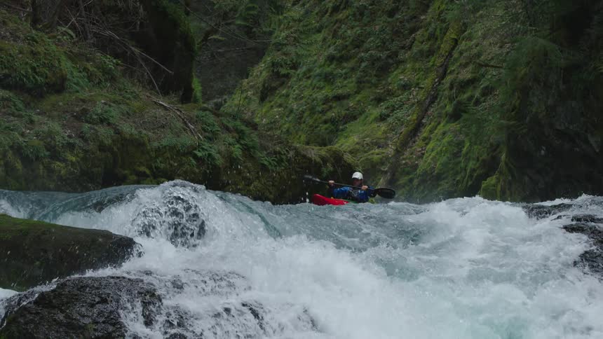 Slow motion shot of a man in red whitewater kayak paddling through an impossibly beautiful mossy canyon towards a steep and dramatic whitewater rapid