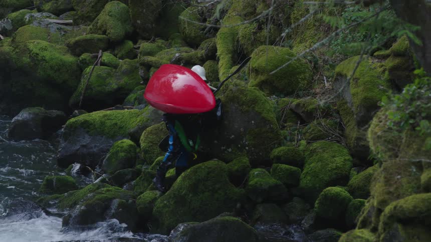 A young adventurer hikes up mossy green rocks on the side of a turbulent river carrying his red whitewater kayak