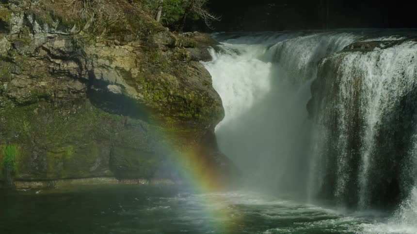 A rainbow shimmers near the rushing splashing waterfall deep in the heart of the wilderness as the camera pans left to right