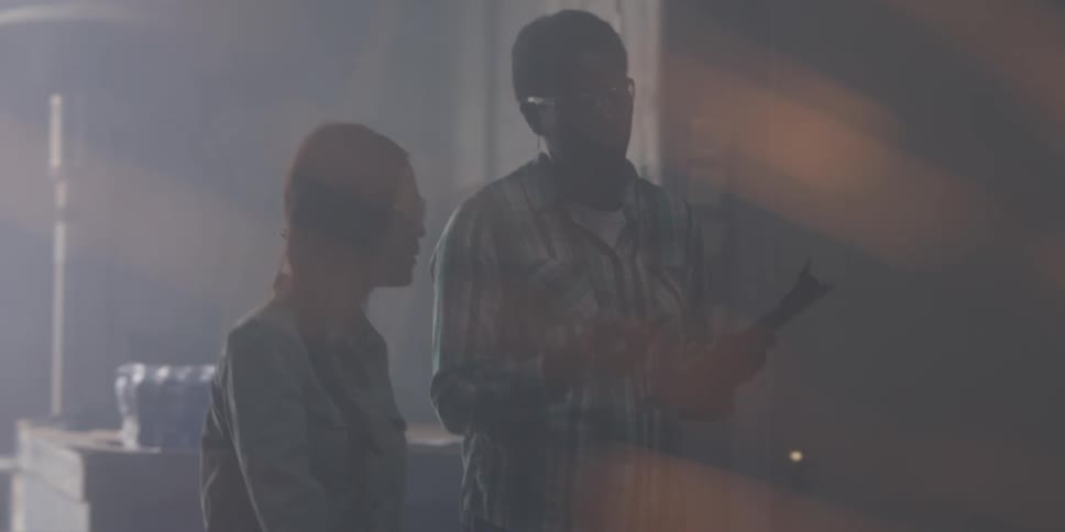 A woman holding a clipboard walking next to another woman who listens to her talk inside of a factory where sparks are flying