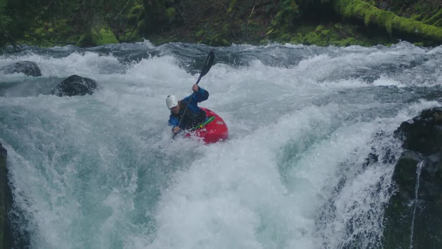 Slow motion shot of a man in a red whitewater kayak wearing a blue drysuit and white helmet riding off a large waterfall and emerging from the pool below
