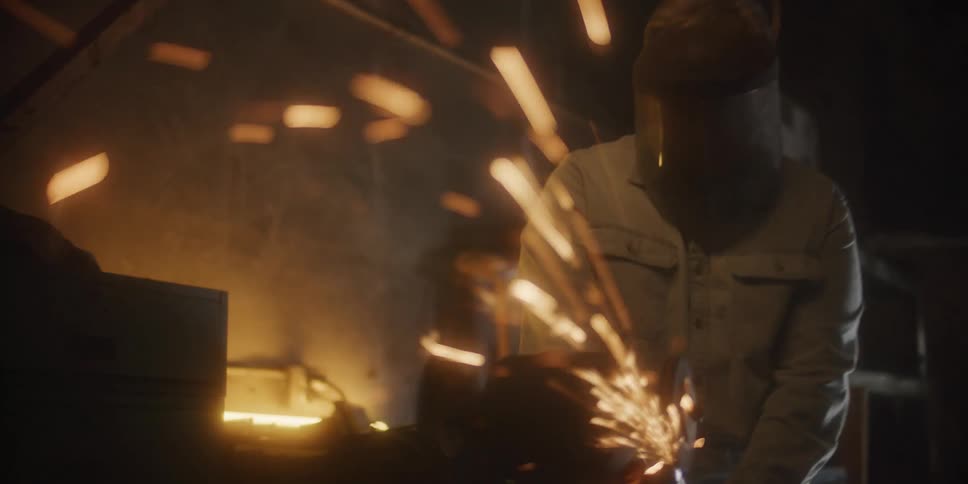 A person with a welding helmet in a workshop sanding metal with sparks with a wall in the background