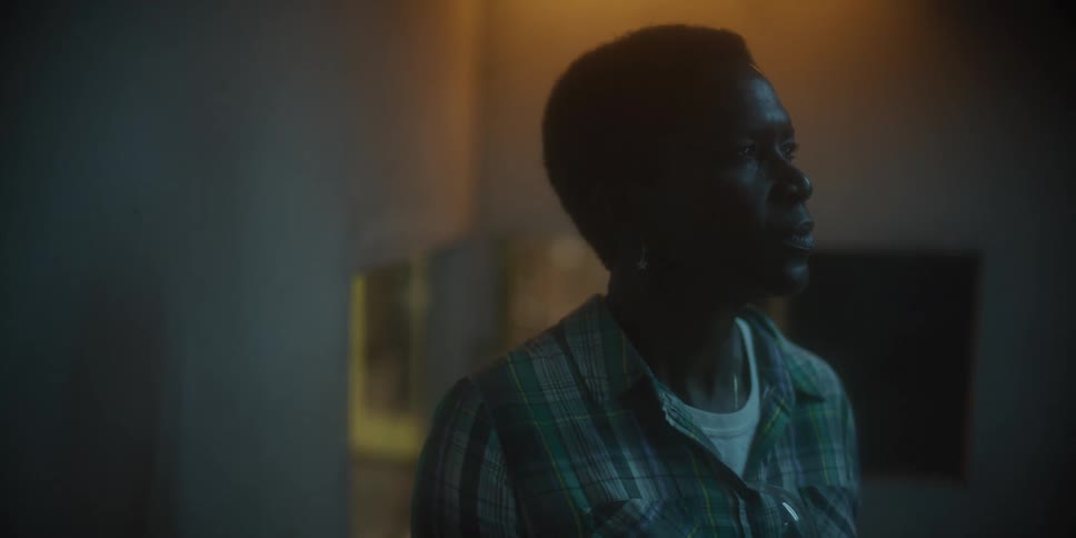 A black man stands looking out with yellow light glowing through window behind him