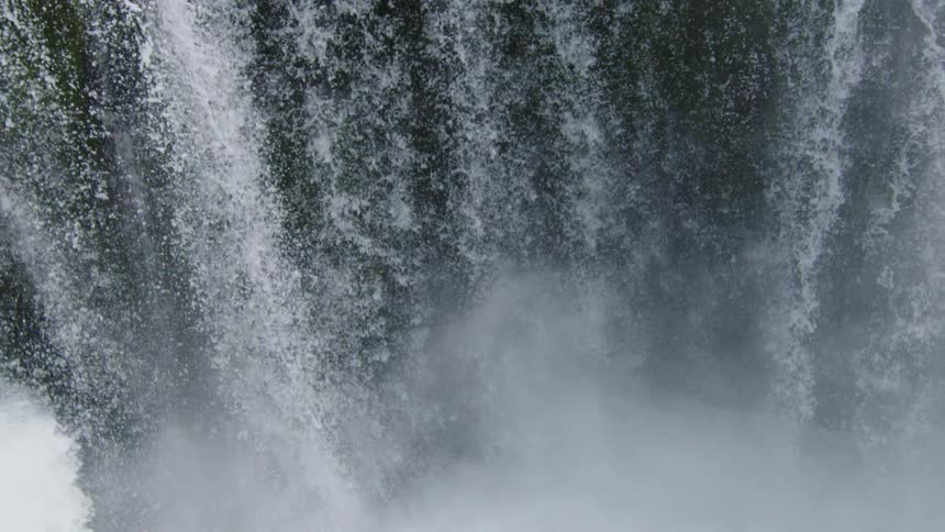Slow motion tilt down of water pouring of the lip of the waterfall before crashing into the narrow river canyon pool below