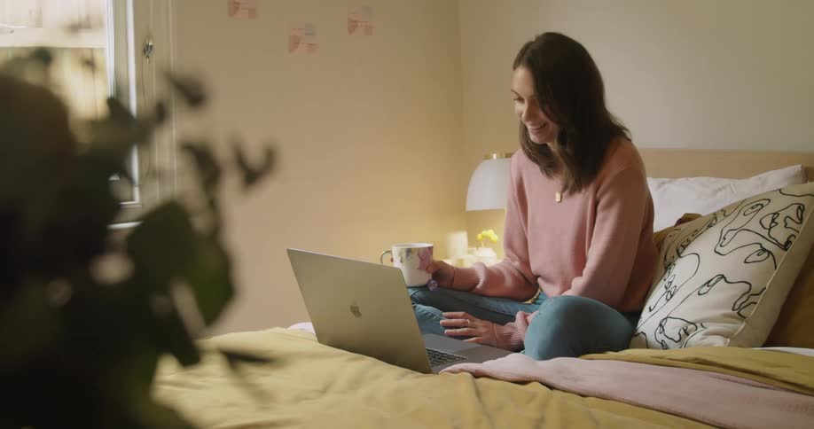 A woman sits on the bed with a mug using her laptop