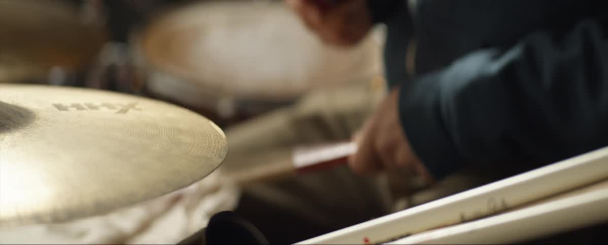 Man with a dark blue zip up hoodie and brown pants plays the drums