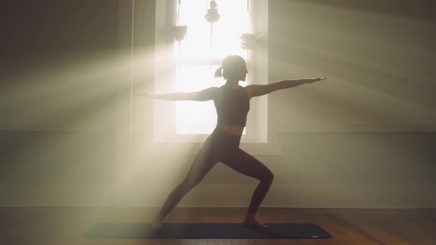 A woman doing yoga in front of a bright sunny window with hanging plants