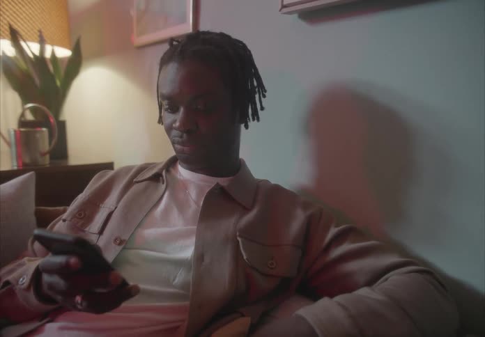 Man with braids and a brown with white shirt sits on the couch and uses his smartphone in the living room at home