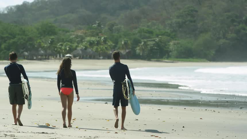 Female and male surfers carrying surfboards walk on the beach next to the sea