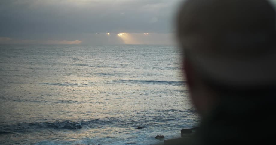 Man stands looking out toward horizon where sun shines through clouds onto ocean