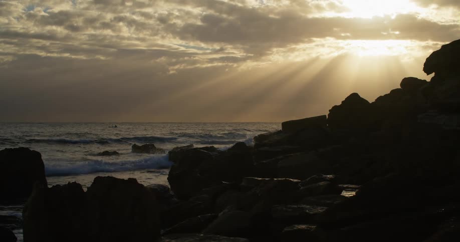 Sunlight shines through gaps in the cloudy sky onto ocean waves rolling into a coastline covered in stones and boulders
