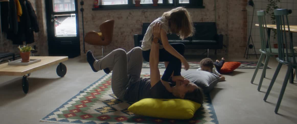 A father lies on the floor at home and lifts his son over his head as his other son watches