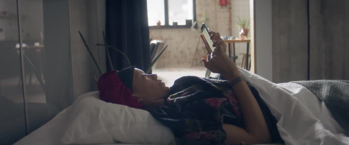 A woman lies in bed at home and looks at her cell phone