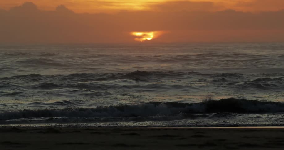 Ocean waves washing onto a beach with an orange sky in the distance at sunset
