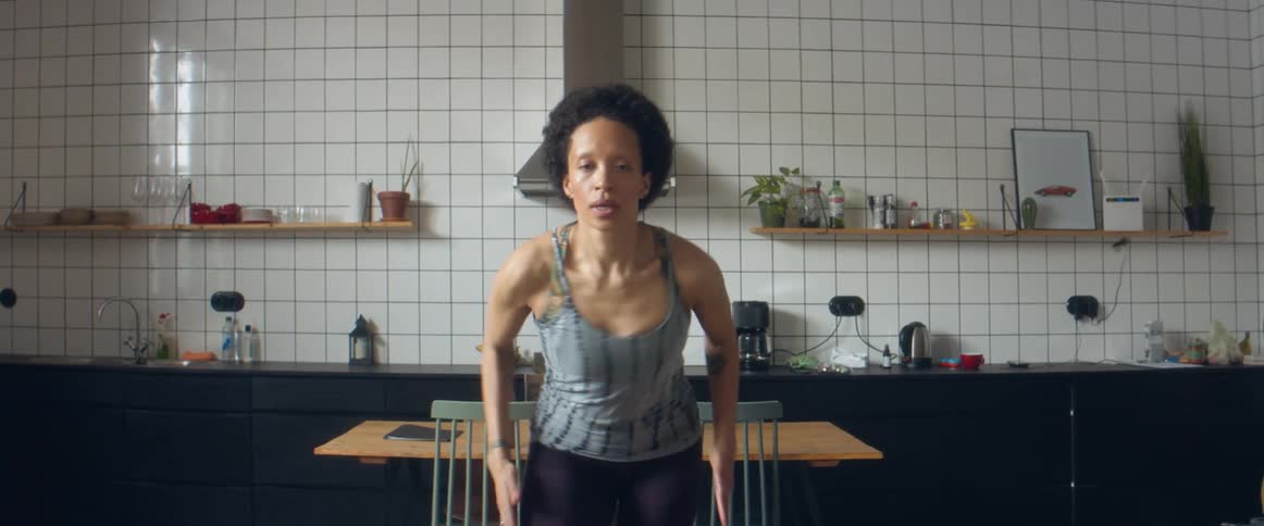 A woman shakes her arms and dances in place as she builds up to a fast run while working out at home in her kitchen