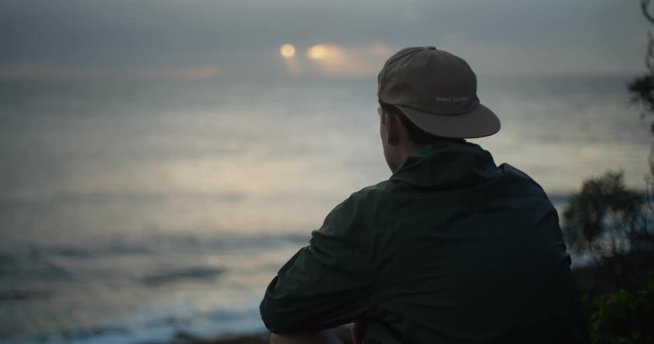 A young man sits on the ground with his hands resting on his knees as he looks at the ocean below