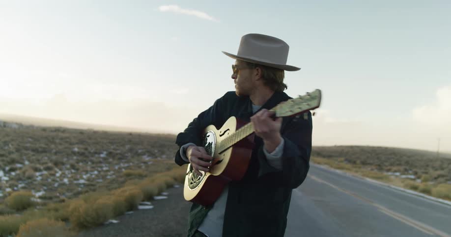 A man walks down a road next to a field playing the guitar and singing