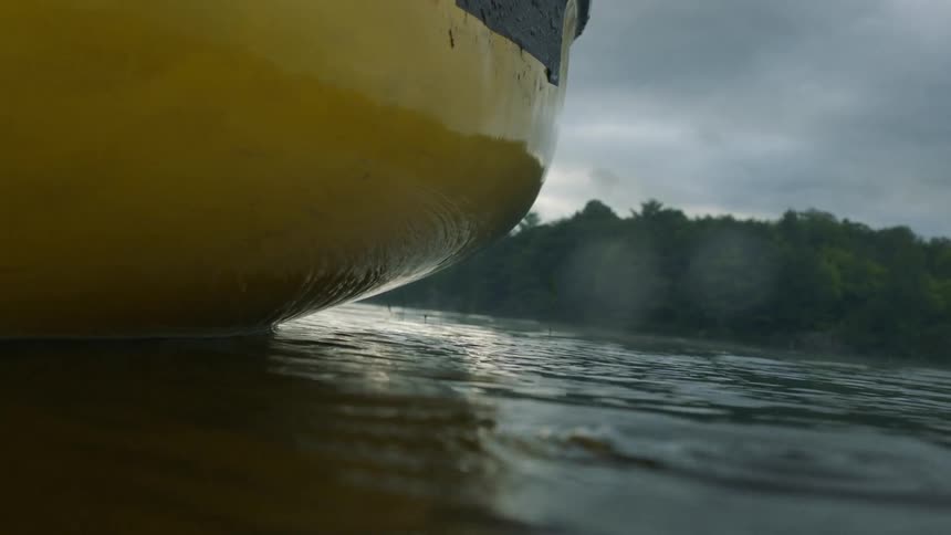 Water drips onto the water surface causing ripples near a yellow boat with tree covered land behind
