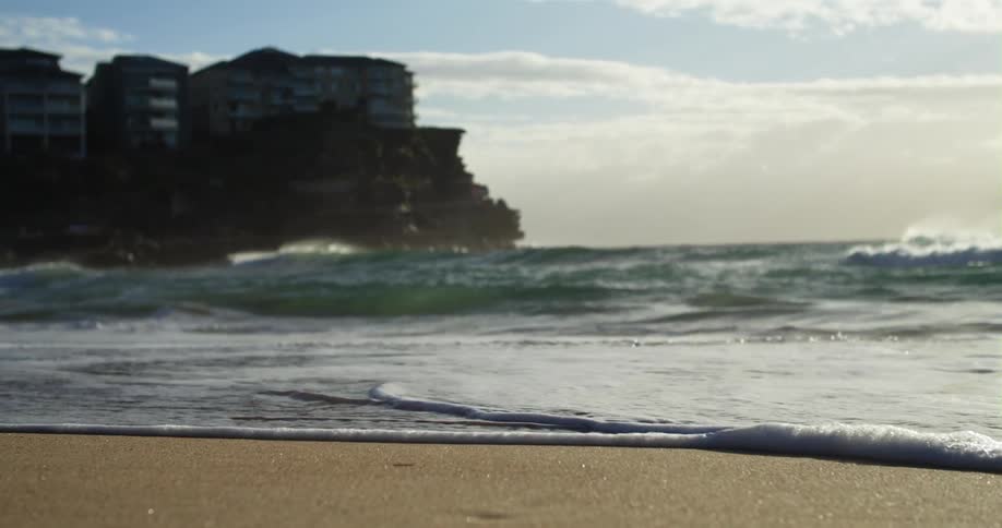 Ocean waves roll into a sandy beach below a cliff holding a large building during the day