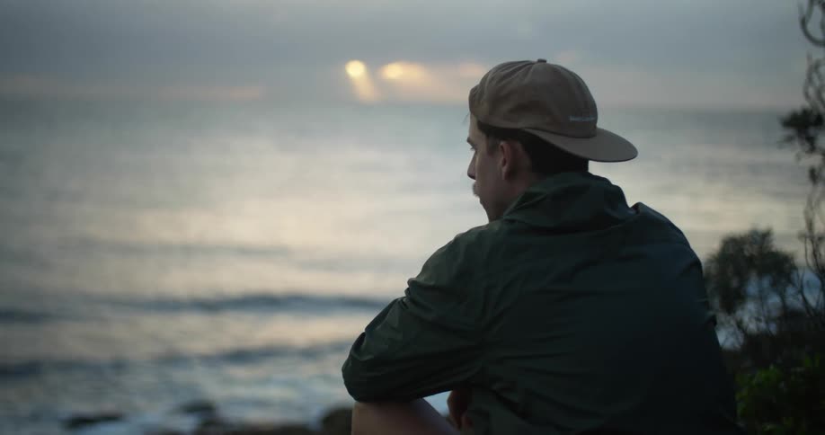Man wearing a baseball hat sitting in front of the ocean at sunset