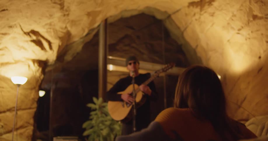 Woman sits on a couch to watch a man playing acoustic guitar on a stage