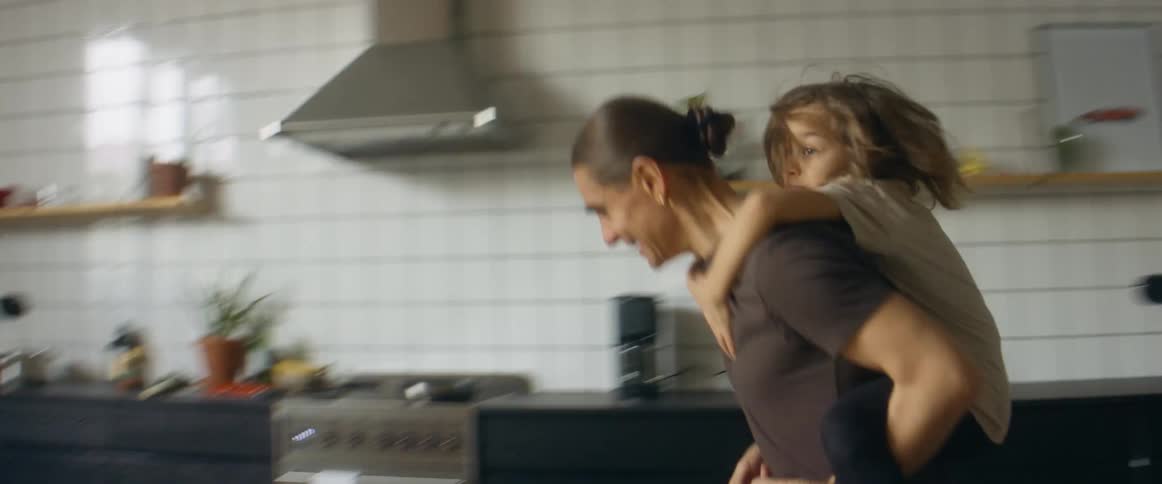 A man laughs as he runs around his house with his son on his back and his wife watches from the couch
