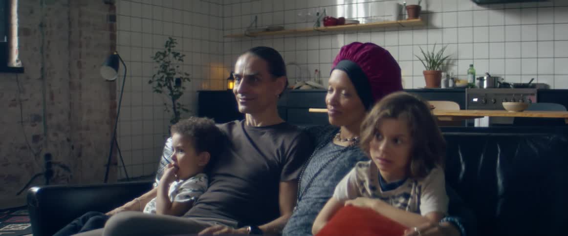 Father and mother look at each other and watch television with their children sitting on the black sofa in the living room at home