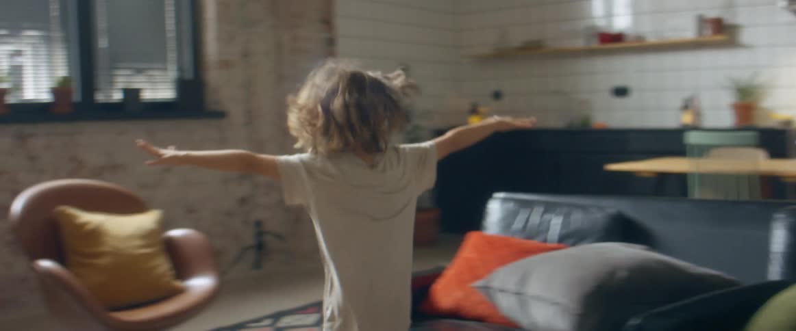 A boy and a girl run around a couch near a kitchen