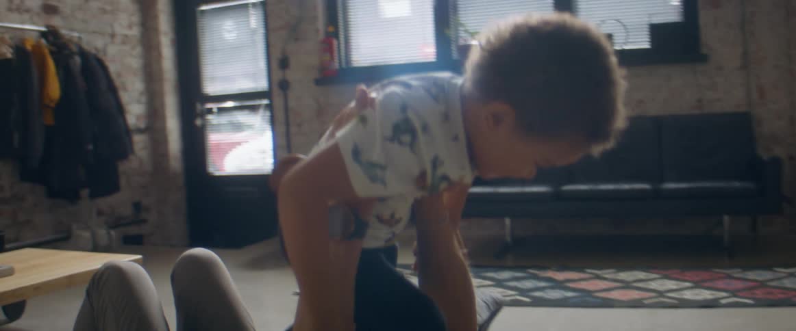 A man lies on the floor at home and holds his son up in the air as his other son sits on the floor with them