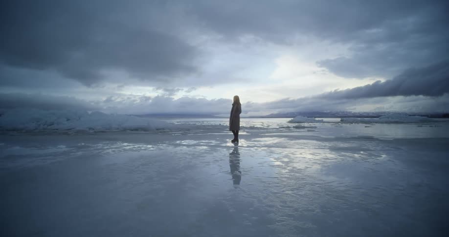 A person in a long coat walking in a desolate cloudy area by a lake